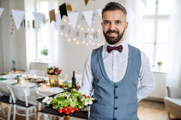 A mature man standing indoors in a room set for a party, holding a tray with vegetables.