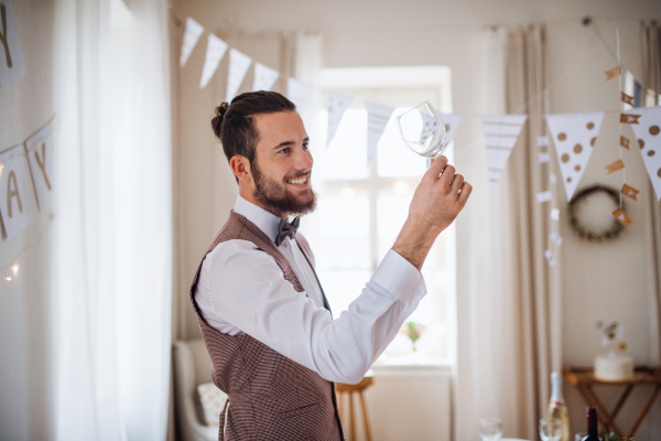 A portrait of a young man indoors in a room set for a party, looking at and checking wine glass.