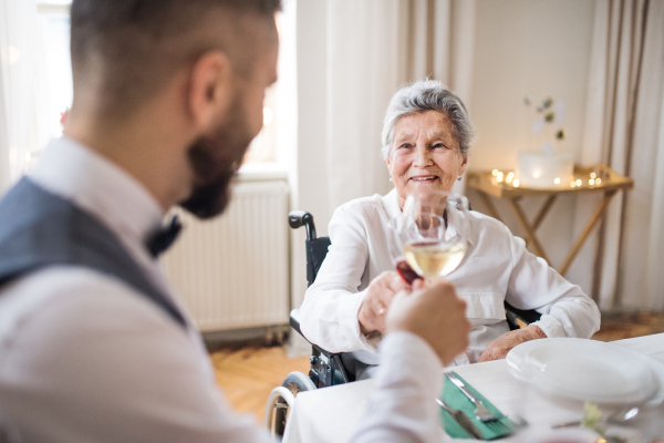 An elderly woman in wheelchair sitting at the table on a indoor birthday party, clinking glasses.