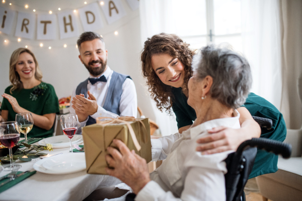 An elderly grandmother in wheelchair celebrating birthday with family and recieving a gift box, a party concept.