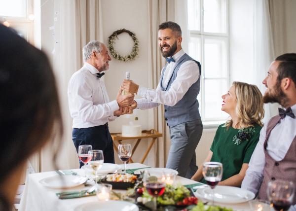 A mature man giving a bottle of wine to his father on indoor birthday party, a celebration concept.