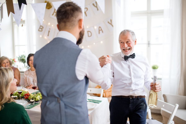 A mature man giving a bottle of wine to his father on indoor birthday party, a celebration concept.