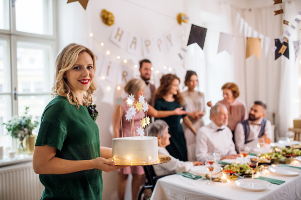 A young woman holding a birthday cake on an indoor party, multigeneration family in the background.