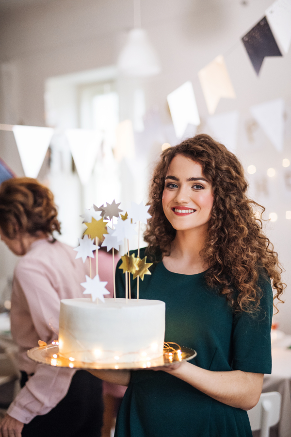 A young woman holding a birthday cake on an indoor party, family in the background.