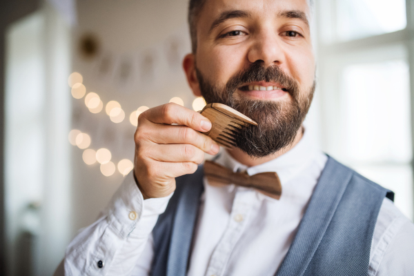 Hipster man standing indoors in a room set for a party, combing beard with a comb.