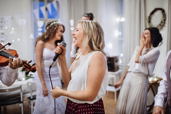 A young woman singing on a wedding reception, bride and guests dancing in the background.