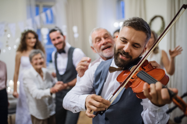 A mature man playing a violin on a wedding reception, bride, groom and other guests dancing.