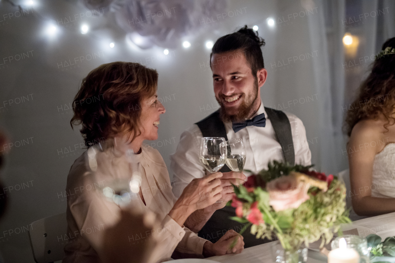 Happy young groom and his mother sitting at a table on a wedding, clinking glasses.