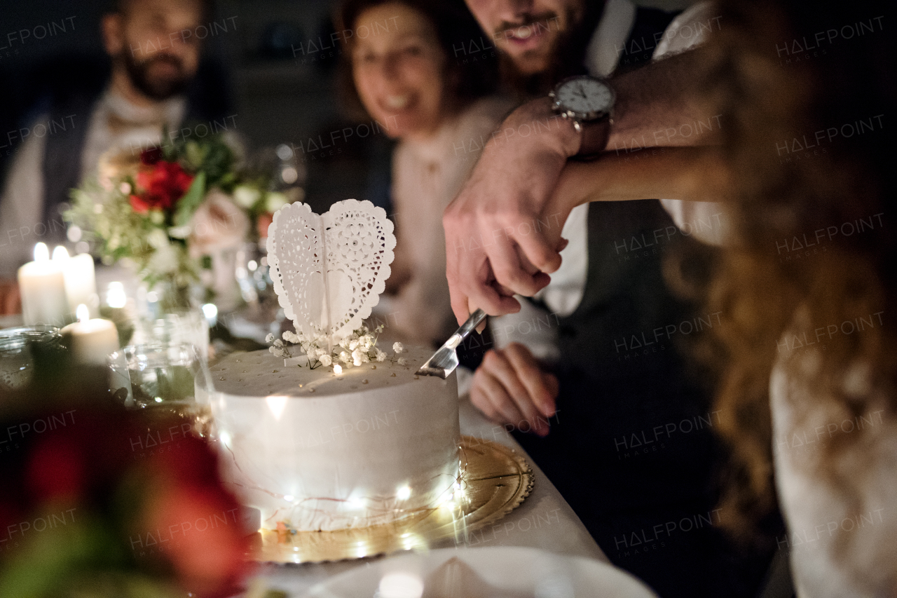 A midsection view of young happy couple sitting at a table on a wedding, cutting a cake.