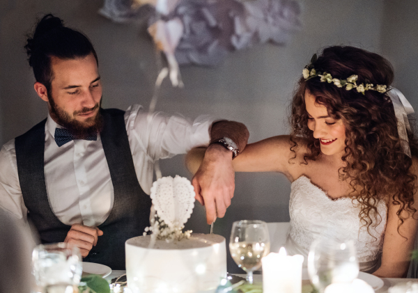 A young happy couple sitting at a table on a wedding, cutting a cake.