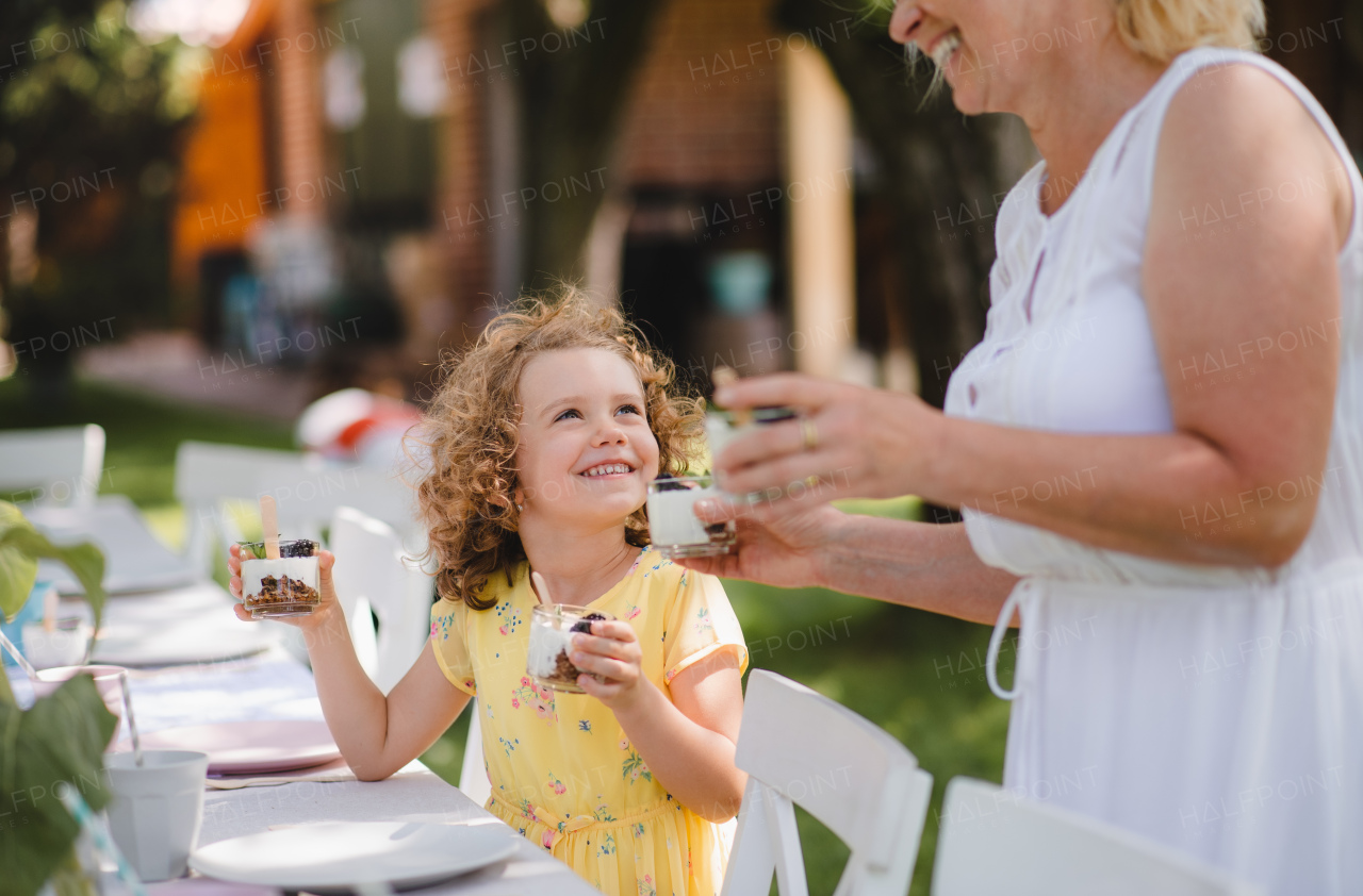 Happy small girl with unrecognizable grandmother outdoors on garden party in summer, eating.