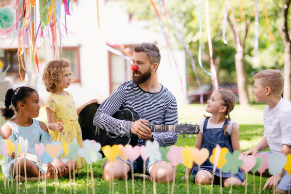 A man with small children sitting on ground outdoors in garden in summer, playing guitar.