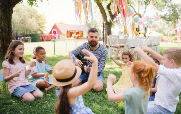 A man with small children sitting on ground outdoors in garden in summer, playing guitar.