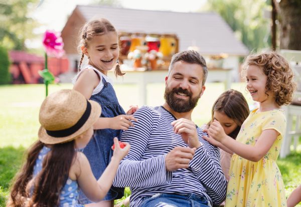 A man with small children sitting on ground outdoors in garden in summer, playing.