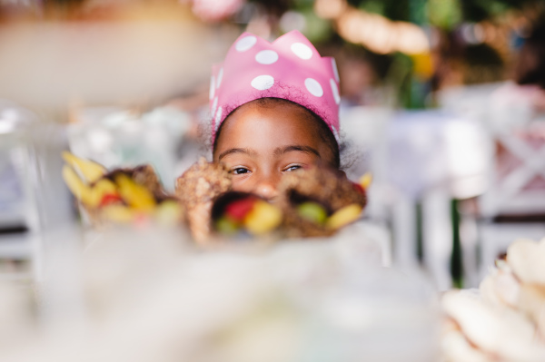 Close-up of small girl with hat outdoors on summer garden party, looking at camera.