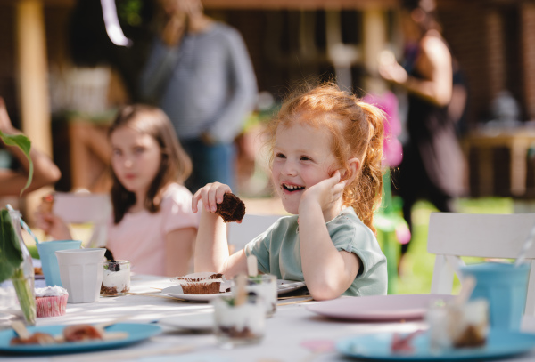 Happy small girls sitting and eating at table on summer garden party, birthday celebration concept.