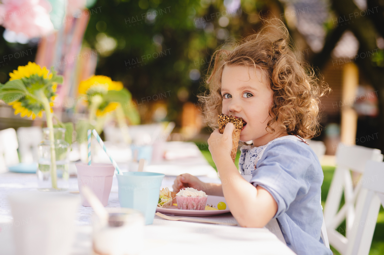 Small girl sitting outdoors in garden in summer, eating snacks. A celebration concept.