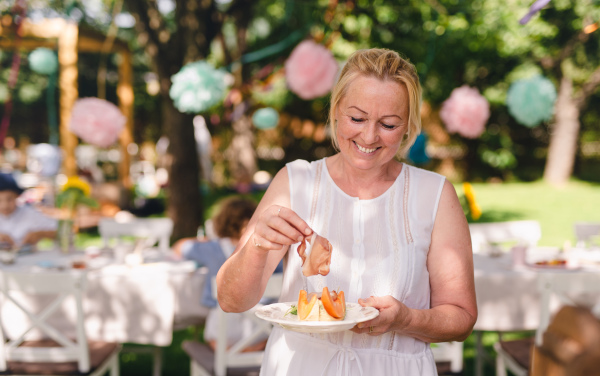 Front view of senior woman standing outdoors on garden party in summer, eating.