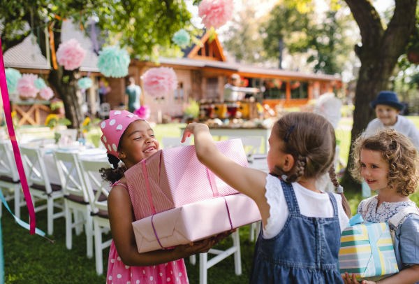 A portrait of small girl with friends and presents outdoors in garden in summer.