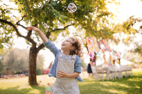 Portrait of happy small girl playing with bubbles outdoors on garden party in summer.