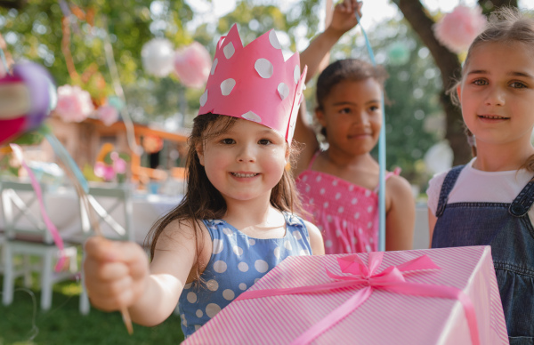 A portrait of small girl with friends and present outdoors in garden in summer.