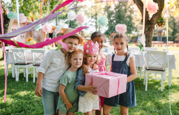 A portrait of small girl with friends and present outdoors in garden in summer.