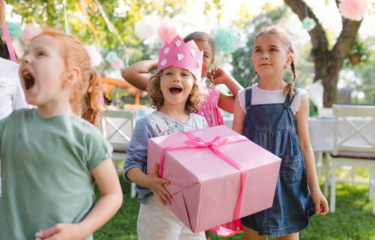 A portrait of small girl with friends and present outdoors in garden in summer.