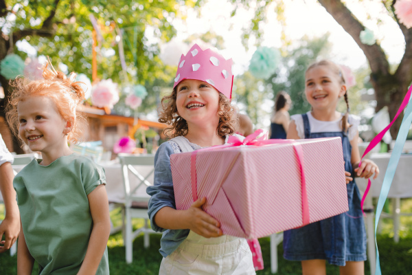 A portrait of small girl with friends and present outdoors in garden in summer.