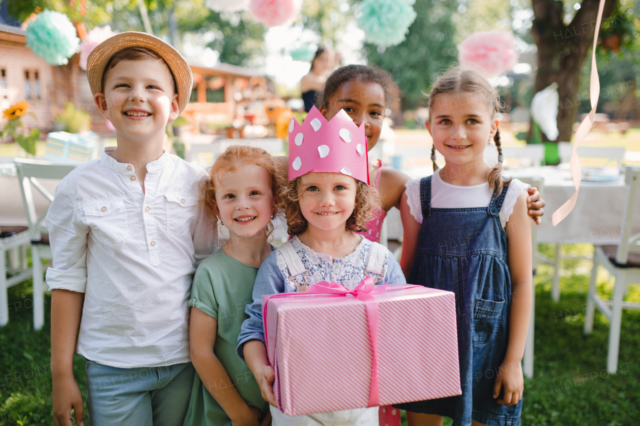 A portrait of small girl with friends and present outdoors in garden in summer.