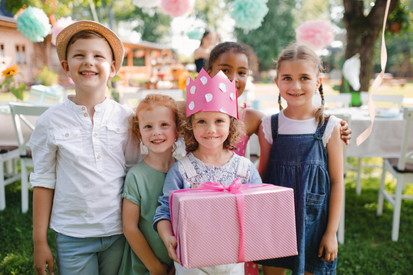 A portrait of small girl with friends and present outdoors in garden in summer.