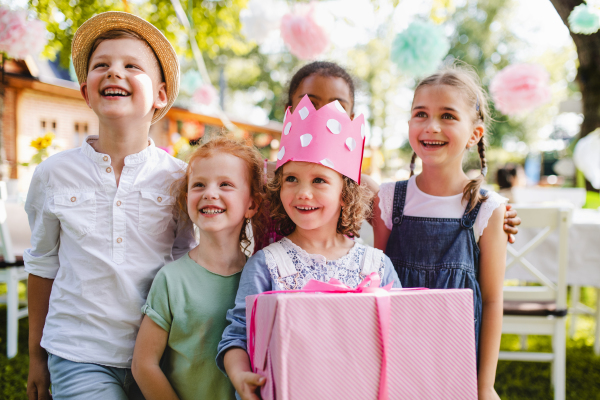 A portrait of small girl with friends and present outdoors in garden in summer.