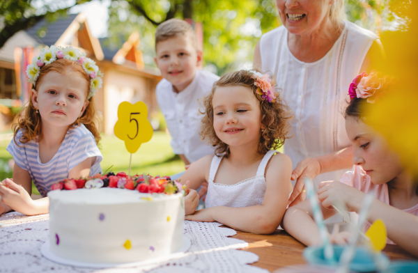 A portrait of children with cake standing around table on birthday party in garden in summer.