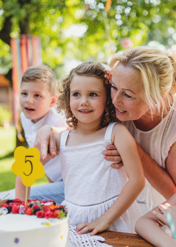 Front view of small girl with cake and grandmother celebrating birthday outdoors in garden in summer, party concept.