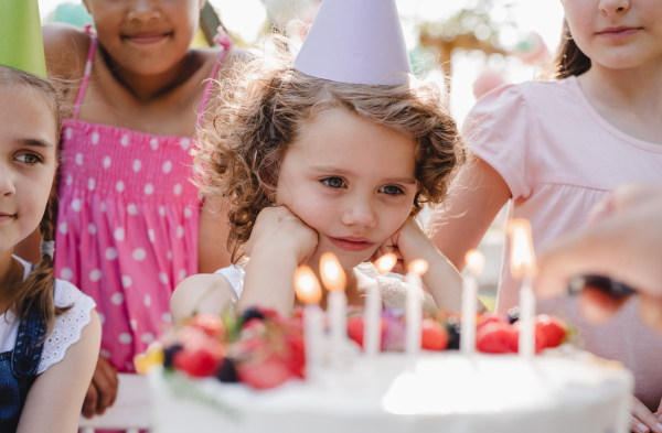 A portrait of children with cake standing around table on birthday party in garden in summer.