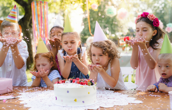 A portrait of children with cake standing around table on birthday party in garden in summer.