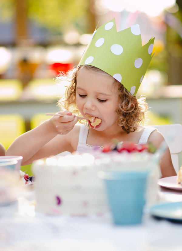 Small girl with party hat sitting outdoors in garden in summer, eating cake. A celebration concept.