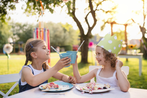 Happy small girls sitting and eating at table on summer garden party, birthday celebration concept.