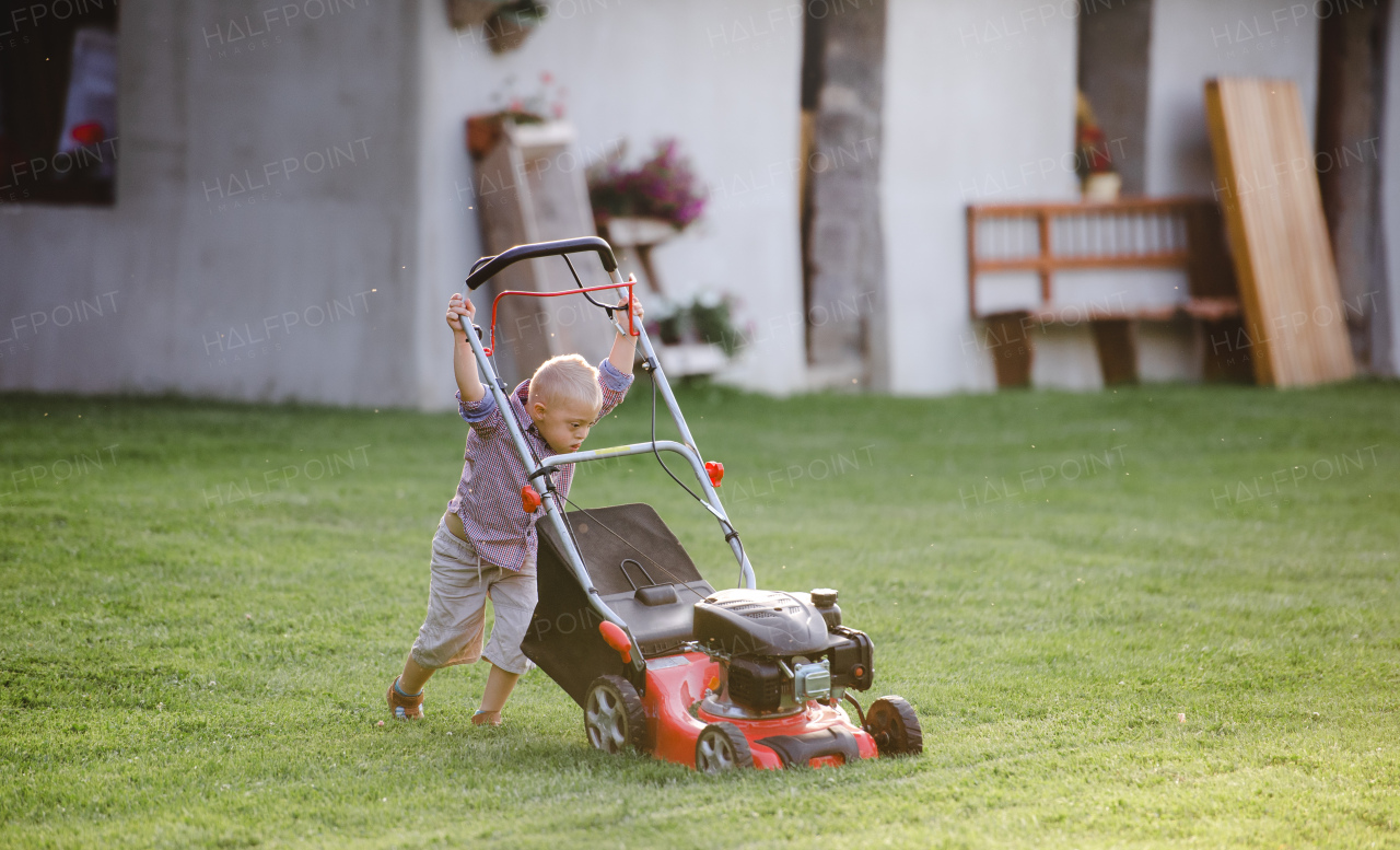 Down syndrome child with lawn mower walking outdoors in garden in summer.