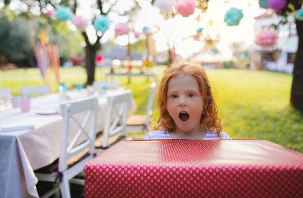 Portrait of small girl with large present box outdoors in garden in summer, looking at camera.