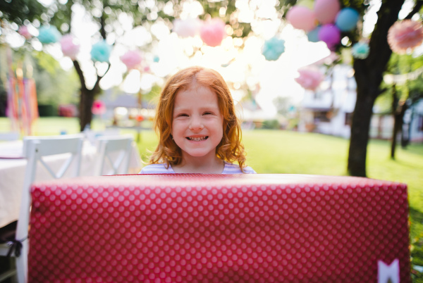 A portrait of small girl with present standing outdoors in garden in summer.