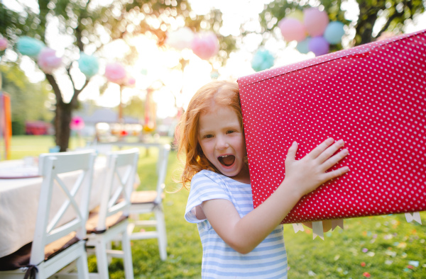 Portrait of small girl with large present box outdoors in garden in summer, looking at camera.