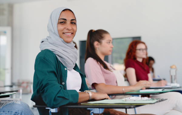 A portrait of islamic university student sitting in classroom indoors, looking at camera.