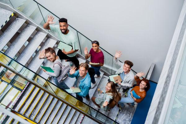 A top view of university students walking up the stairs indoors, looking at camera and waving.