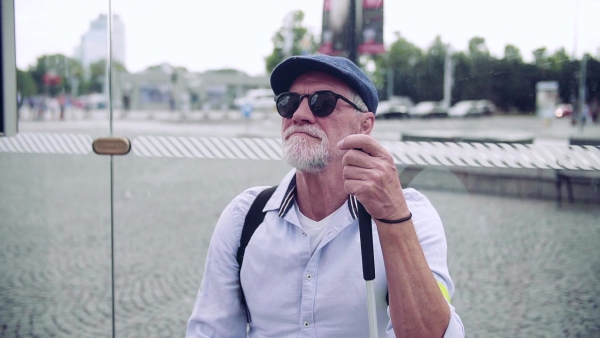 A senior blind man with white cane waiting at bus stop in city. Slow motion.