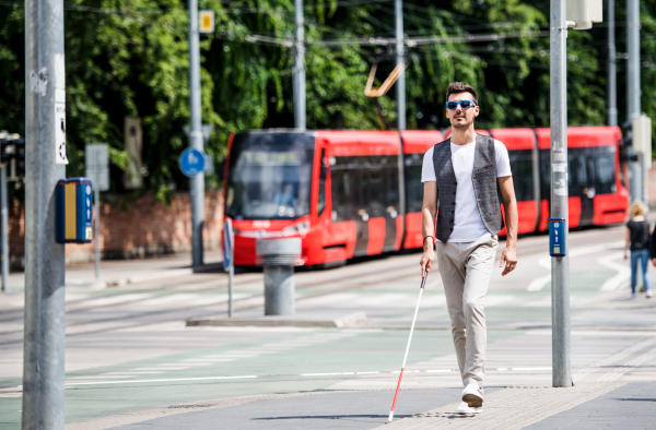 Young blind man with white cane walking across the street in city. Copy space.