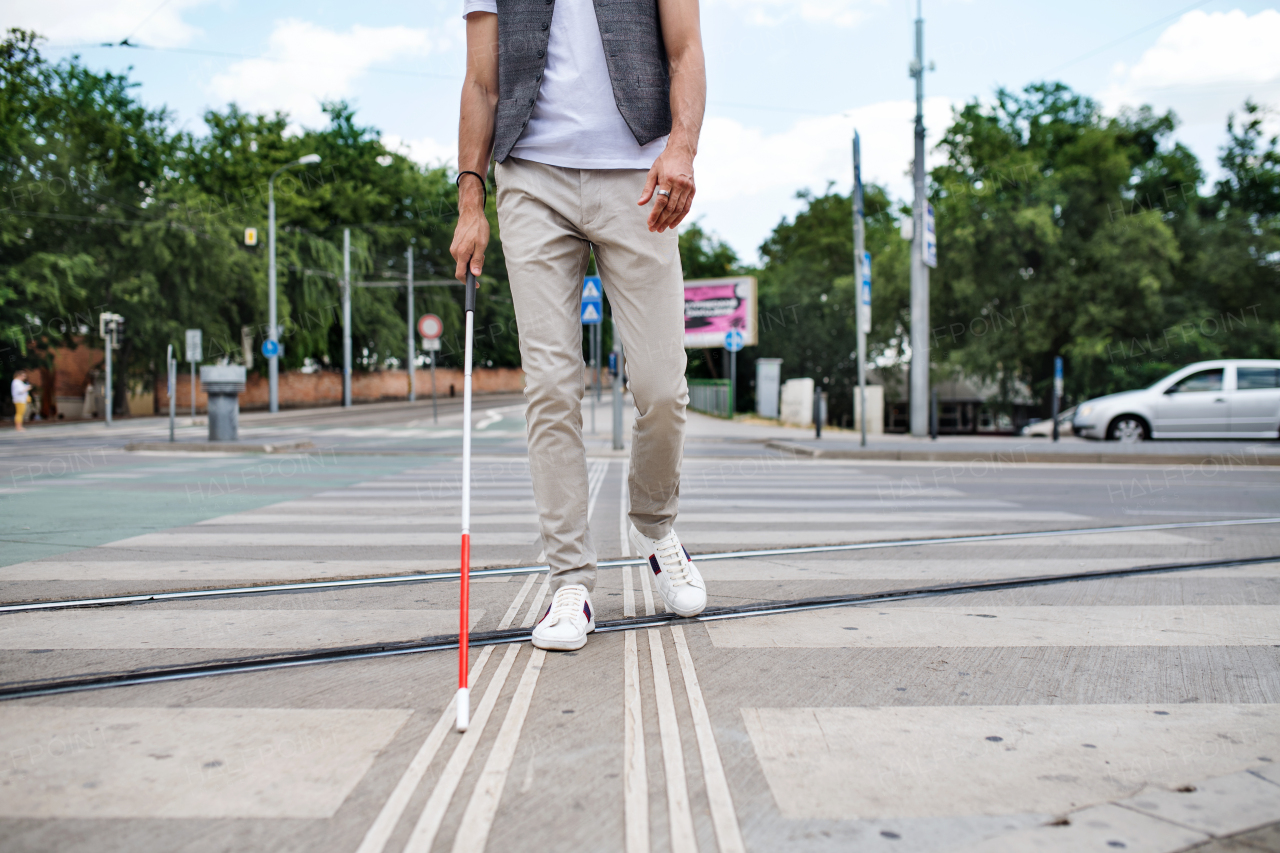 Midsection of young unrecognizable blind man with white cane walking across the street in city.