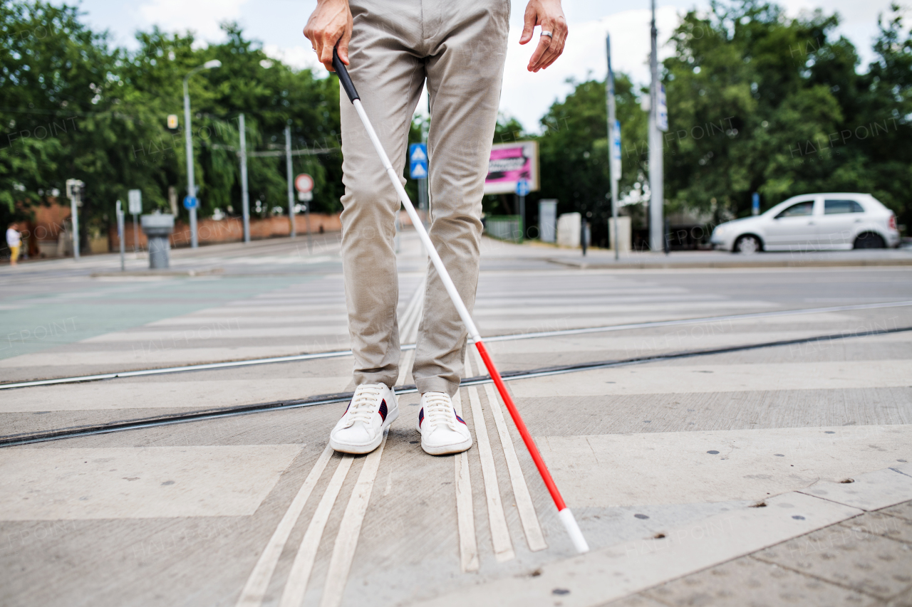 Midsection of young unrecognizable blind man with white cane walking across the street in city.