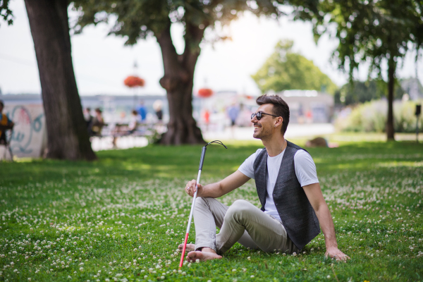 Young blind man with white cane and guide dog sitting in park in city, resting.