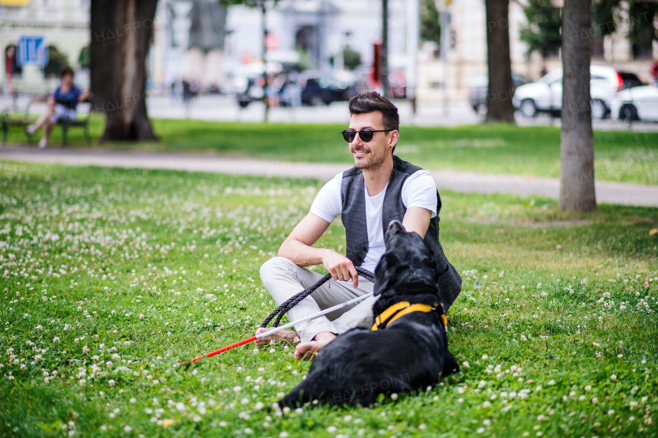 Young blind man with white cane and guide dog sitting in park in city, resting.