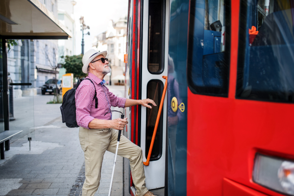 A senior blind man with white cane getting on public transport in city.